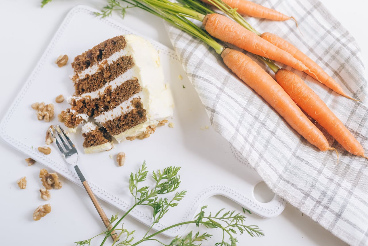 A slice of carrot cake with frosting, alongside fresh carrots. Perfect for dessert lovers.